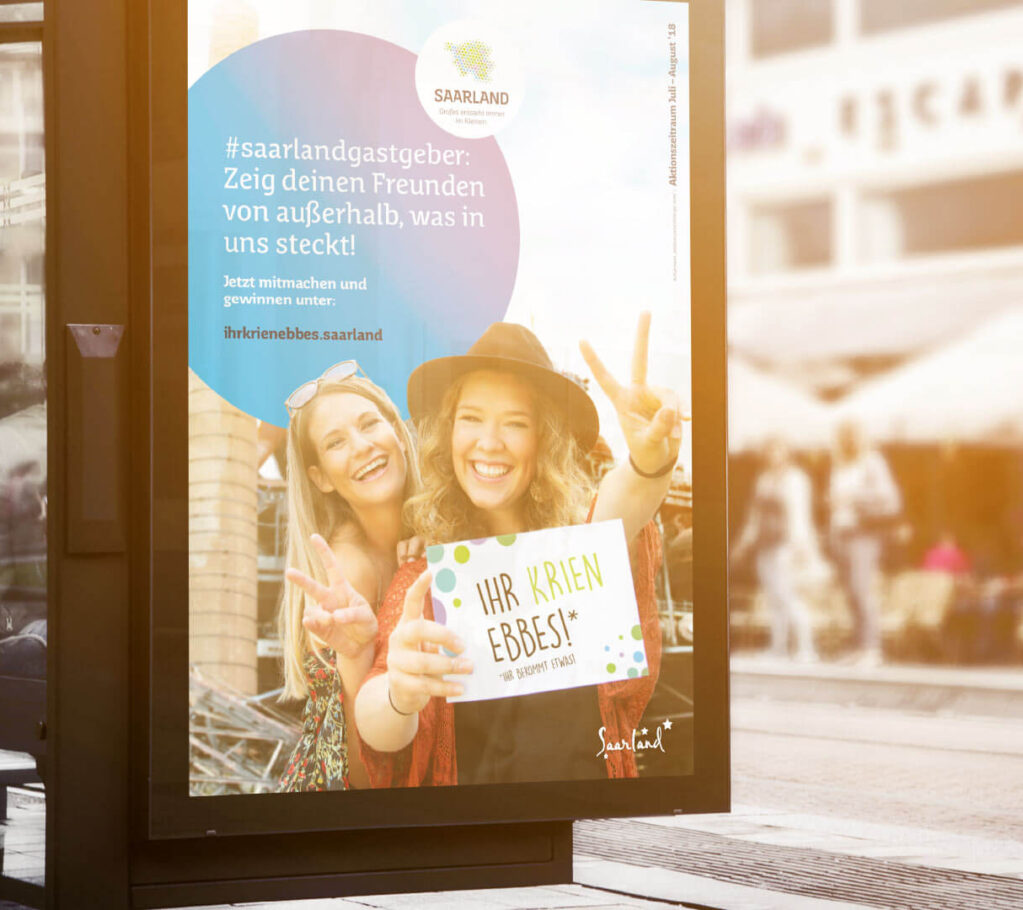 Ein Plakat an einer Bushaltestelle zeigt zwei lächelnde Frauen, die ein Schild mit der Aufschrift „Ihr krien Ebbes“! halten. Die Mitmachkampagne wirbt für den saarländischen Tourismus mit Hashtags und einer Botschaft, die Freunde zum Mitmachen ermutigt – Ihr krien ebbes wartet!.