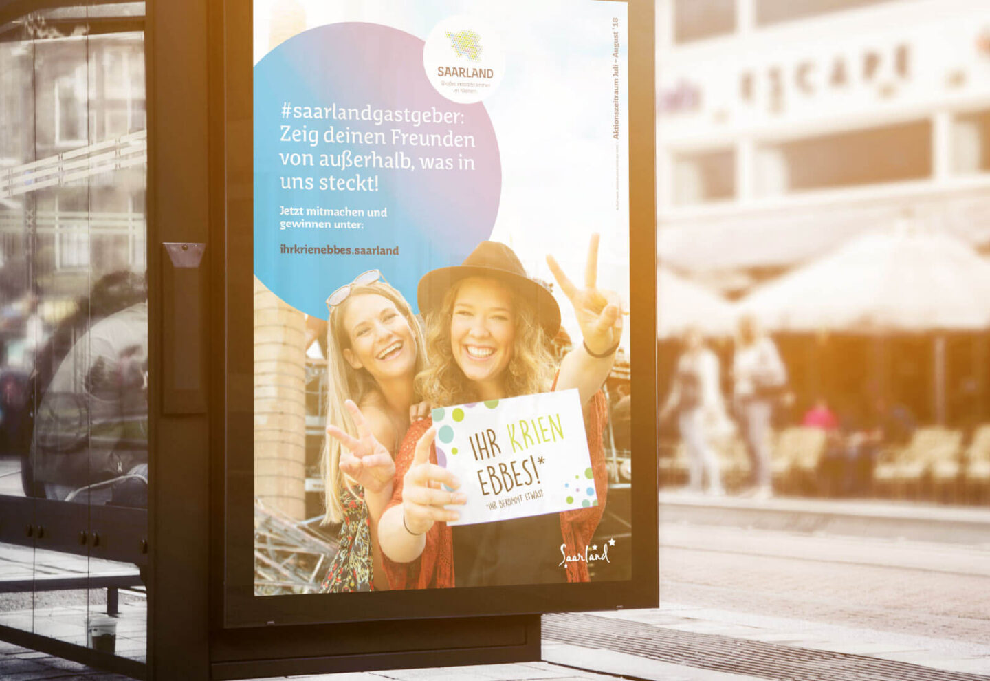 Ein Plakat an einer Bushaltestelle zeigt zwei lächelnde Frauen, die ein Schild mit der Aufschrift „Ihr krien Ebbes“! halten. Die Mitmachkampagne wirbt für den saarländischen Tourismus mit Hashtags und einer Botschaft, die Freunde zum Mitmachen ermutigt – Ihr krien ebbes wartet!.