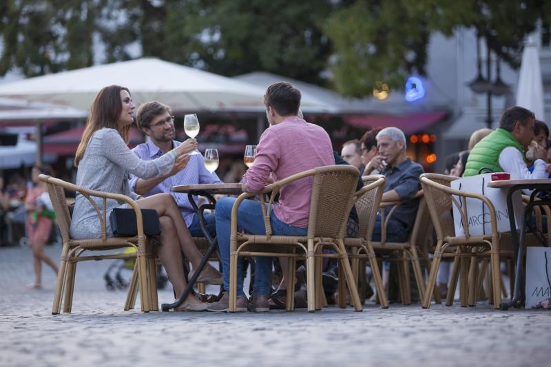 Drei Personen sitzen an einem Cafétisch im Freien auf einer Kopfsteinpflasterstraße im Saarland, Deutschlands Hidden Champion, halten Gläser in der Hand und unterhalten sich. Andere Gäste sitzen in der Nähe, mit großen Sonnenschirmen und Bäumen im Hintergrund - eine einladende Szene für Saarland Marketing.