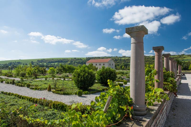 Ein Landschaftsgarten mit gestutzten Sträuchern, Kieswegen und hohen Steinsäulen unter einem blauen Himmel. Im Hintergrund steht ein weißes Gebäude mit rotem Dach - ein Beispiel für das Standortmarketing Saarland in der Region Hidden Champion in Deutschland.