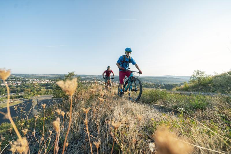 Zwei Personen fahren Mountainbike auf einem grasbewachsenen Hügel im Saarland, mit Blick auf eine entfernte Stadt und einen klaren Himmel - ein Outdoor-Abenteuer, das von Saarland Marketing perfekt eingefangen wurde, um Deutschlands Hidden Champion zu präsentieren. Vertrocknete Pflanzen zieren das unebene Gelände im Vordergrund.