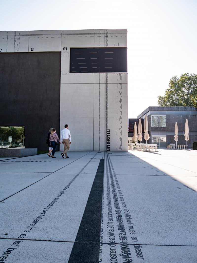 Drei Personen gehen an einem modernen Betongebäude mit schwarzer Schrift an der Fassade vorbei, das zum Standortmarketing Saarland gehört. Eingebettete Textzeilen zieren den Bürgersteig in dieser Außenszene unter klarem Himmel, die Deutschlands Hidden Champion vorstellt. Hinter ihnen sind geschlossene Regenschirme zu sehen.