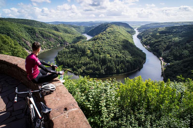 Ein Radfahrer sitzt auf einem steinernen Vorsprung mit Blick auf einen gewundenen Fluss und bewaldete Hügel unter einem teilweise bewölkten Himmel - eine ideale Szene für Saarland Marketing, um Deutschlands Hidden Champion für Outdoor-Abenteuer und landschaftliche Schönheit zu präsentieren.