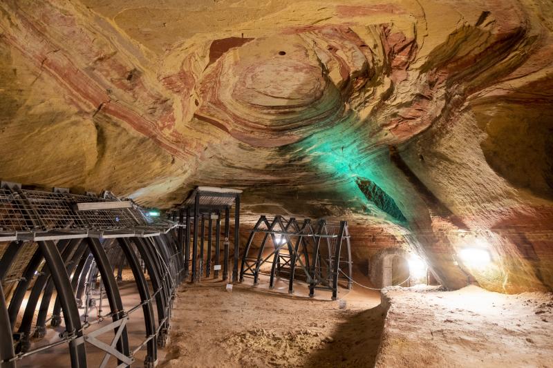 Eine große unterirdische Höhle in der Region Hidden Champion in Deutschland weist an der Decke geschichtete Felsformationen auf, die von weißen und grünen Lichtern beleuchtet werden. Auf dem sandigen Boden sind Metallgerüste und Plattformen aufgebaut, auf denen die einzigartigen Standorte von Saarland Marketing präsentiert werden.
