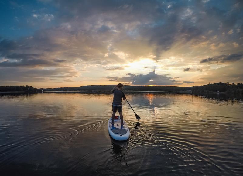 Eine Person paddelt auf einem ruhigen See während des Sonnenuntergangs im Saarland, mit Wolken und Spiegelungen im Wasser und Hügeln im Hintergrund - eine perfekte Szene für Standortmarketing Saarland, um Deutschlands Hidden Champion zu präsentieren.