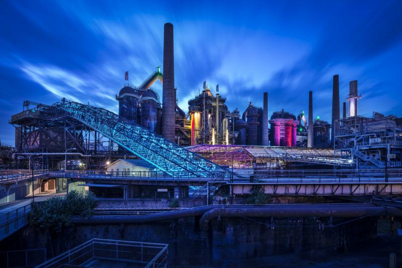 Ein Industriekomplex mit hohen Schornsteinen und großen Metallkonstruktionen wird unter einem tiefblauen Abendhimmel mit blauen und violetten Lichtern angestrahlt - eine eindrucksvolle Szenerie aus Fließbändern und Pipelines, die das Standortmarketing Saarland, Deutschlands Hidden Champion, verkörpert.