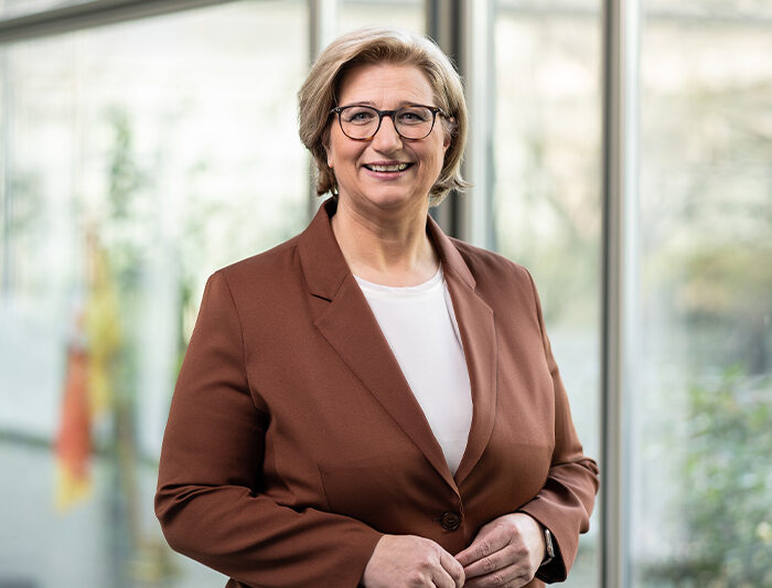 Eine Frau mittleren Alters mit kurzen blonden Haaren und Brille, in braunem Blazer und weißem Hemd, steht in einem Innenraum vor großen Fenstern und lächelt in die Kamera - das sympathische Gesicht des Standortmarketings Saarland.