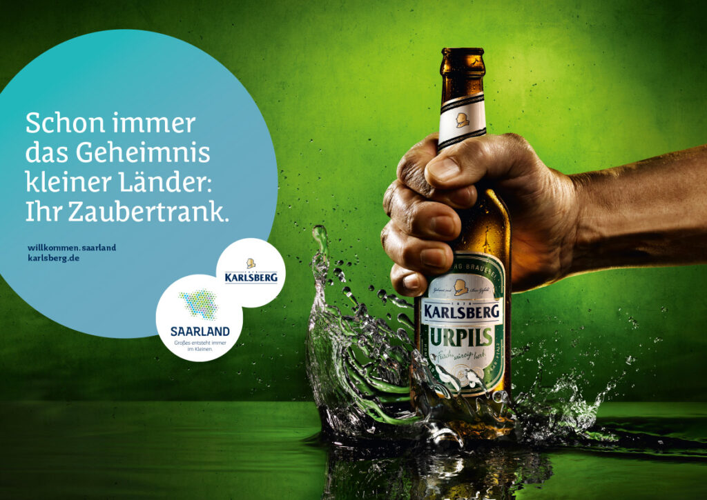 Eine Hand hält eine Karlsberg Urpils-Bierflasche, die vor einem grünen Hintergrund ins Wasser spritzt. Der weiße Text auf der linken Seite lautet: Schon immer das Geheimnis kleiner Länder: Ihr Zaubertrank. Mit Saarland Marketing und zwei Logos am unteren Rand.