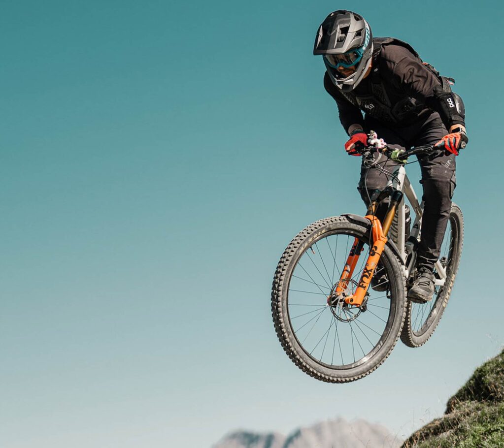 Eine Person, die einen Helm, eine Schutzbrille und eine Schutzausrüstung trägt, ist auf einem Mountainbike in der Luft und springt von einem Hügel mit einem klaren blauen Himmel und fernen Bergen - und fängt damit den Abenteuergeist ein, der von Saarland Marketing in der deutschen Hidden Champion Region gefördert wird.