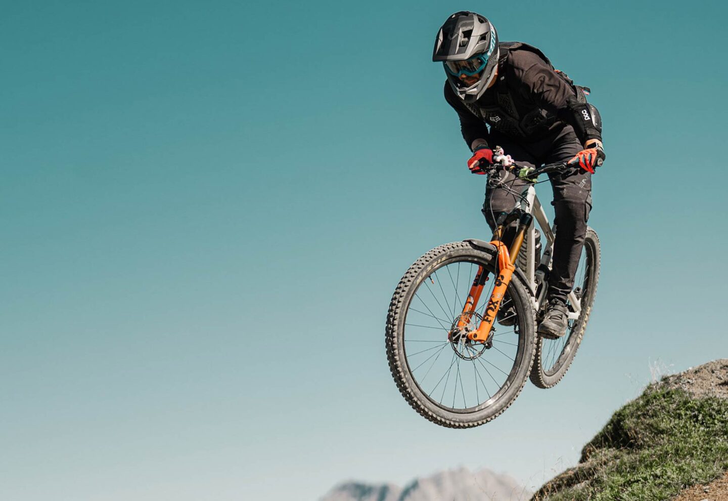 Eine Person, die einen Helm, eine Schutzbrille und eine Schutzausrüstung trägt, ist auf einem Mountainbike in der Luft und springt von einem Hügel mit einem klaren blauen Himmel und fernen Bergen - und fängt damit den Abenteuergeist ein, der von Saarland Marketing in der deutschen Hidden Champion Region gefördert wird.