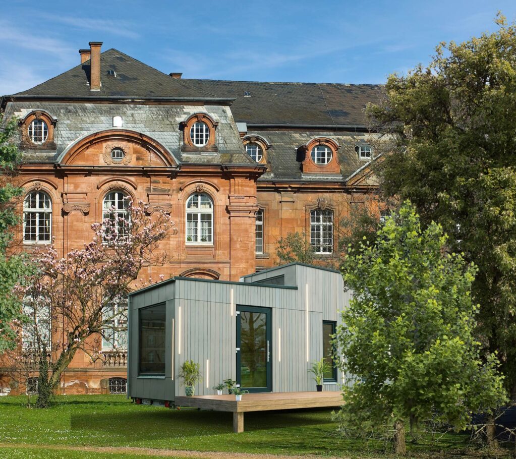 Ein modernes, kastenförmiges, hellgraues Modulhaus steht auf einer Wiese vor einem großen, historischen Backsteingebäude mit Rundbogenfenstern und kunstvollen Details - ein eindrucksvolles Schaufenster des Standortmarketings Saarland. Bäume und Sträucher umgeben beide Gebäude.