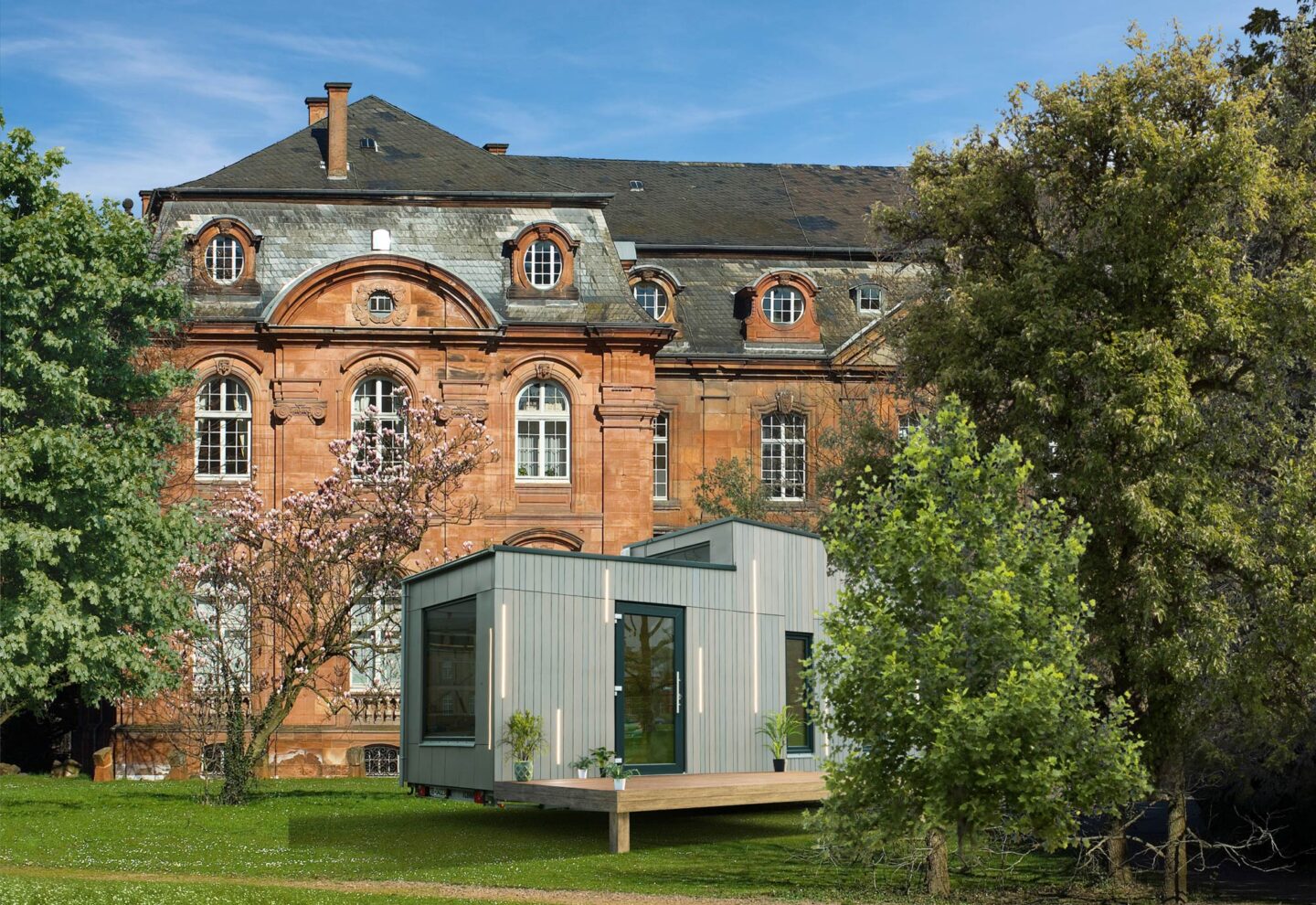 Ein modernes, kastenförmiges, hellgraues Modulhaus steht auf einer Wiese vor einem großen, historischen Backsteingebäude mit Rundbogenfenstern und kunstvollen Details - ein eindrucksvolles Schaufenster des Standortmarketings Saarland. Bäume und Sträucher umgeben beide Gebäude.