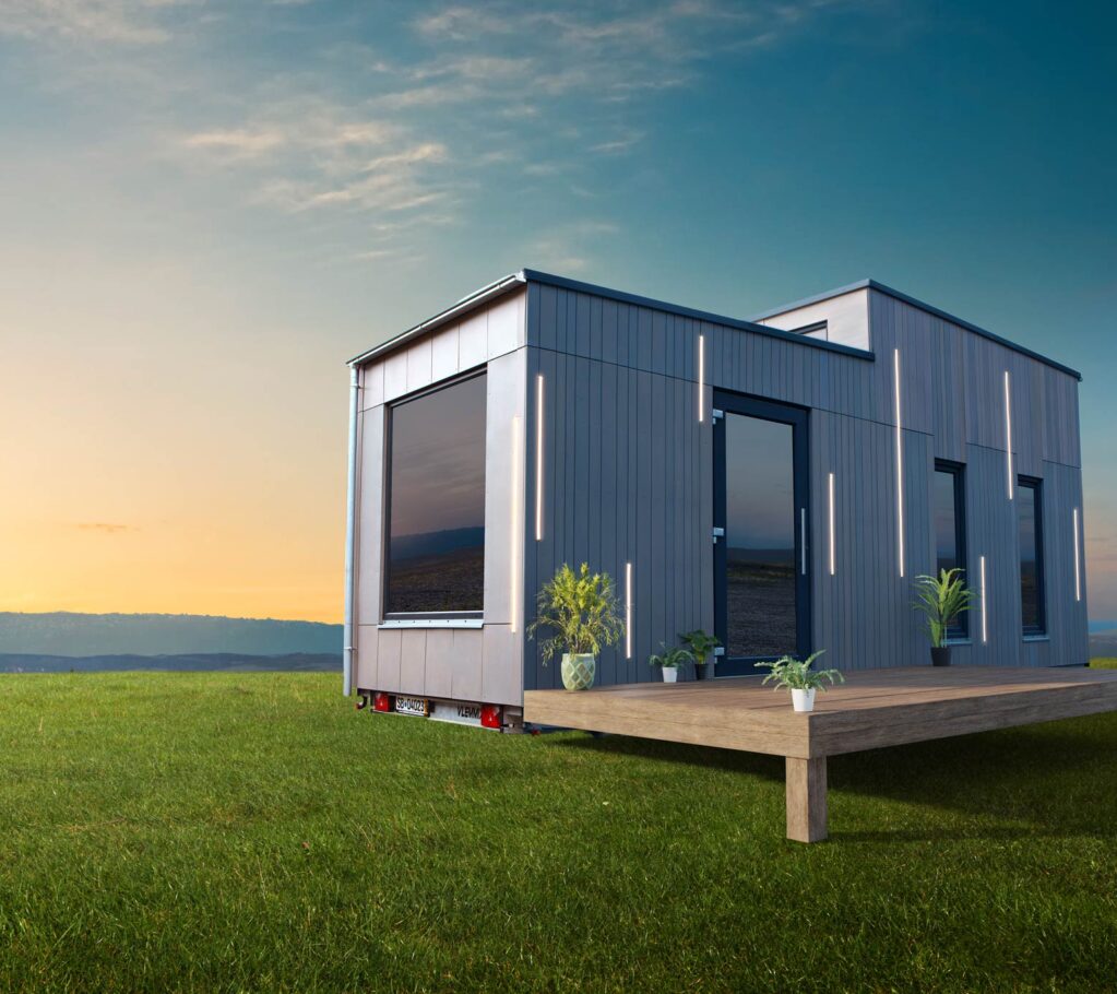 Ein modernes kleines Haus mit großen Fenstern steht bei Sonnenuntergang auf einer Wiese - ein perfektes Beispiel für Saarland Marketing. Davor erstreckt sich eine Holzterrasse mit Topfpflanzen, und der Himmel ist meist klar mit ein paar Wolken.