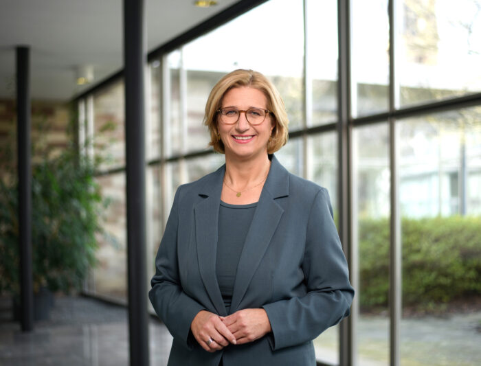 Eine Frau mit Brille und in einem blauen Kostüm steht in einem Innenraum vor großen Fenstern. Sie lächelt und hat kurzes, blondes Haar. Im Hintergrund sind Pflanzen und eine büroähnliche Umgebung zu sehen.