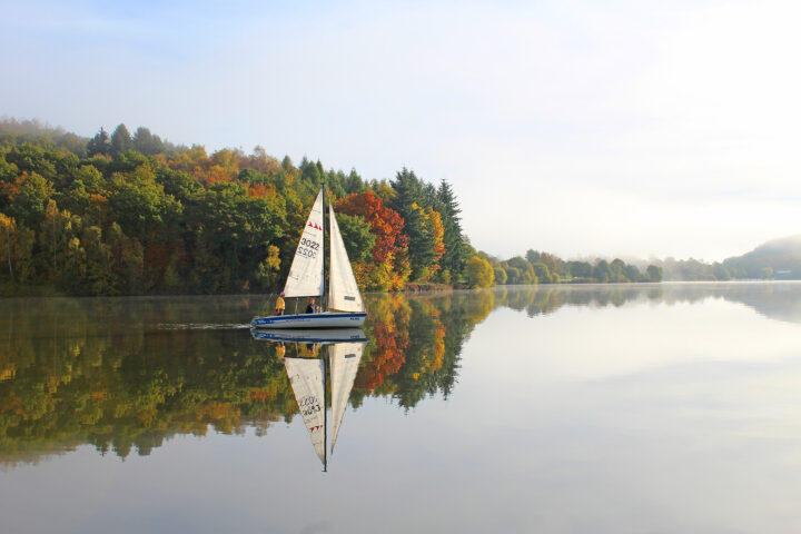 Ein kleines Segelboot gleitet über einen ruhigen See, umgeben von Bäumen mit herbstlichem Laub – eine friedliche Szene, die Teil eines jeden inspirierenden Rahmenprogramms sein könnte. Das Wasser spiegelt das Boot und die farbenprächtigen Bäume perfekt wider, während der Himmel leicht dunstig bleibt und so die malerische Atmosphäre dieses natürlichen Rahmenprogramms 3 unterstreicht.