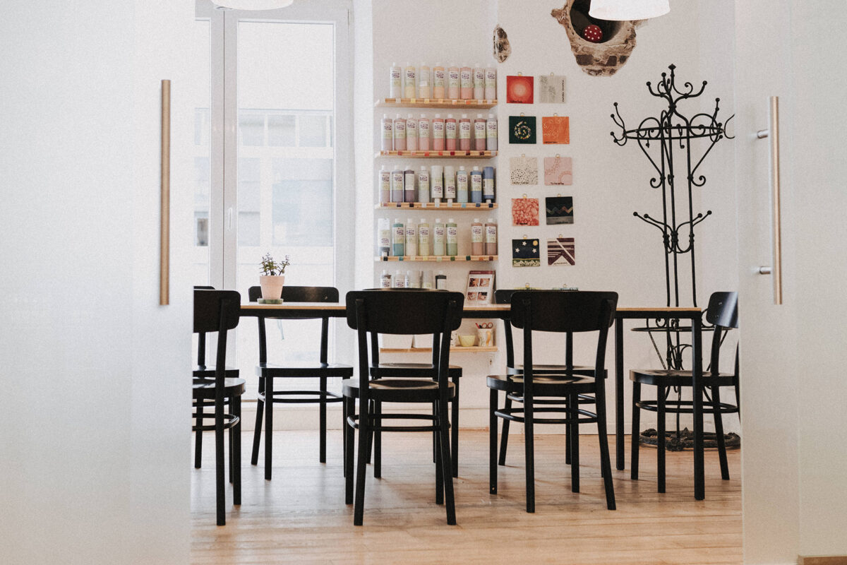 Ein moderner Raum mit einem Holztisch, um den herum schwarze Stühle stehen, einer Garderobe, Wandregalen voller Flaschen, kleinen Wandbildern, die das Rahmenprogramm ergänzen, einer Topfpflanze auf dem Tisch und großen Fenstern, durch die Tageslicht hereinströmt.