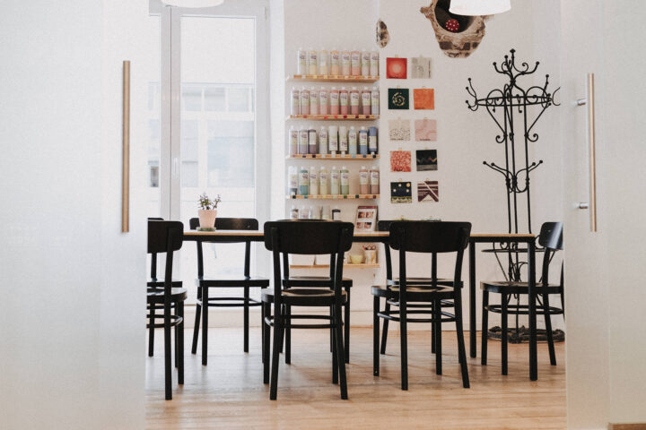 Ein moderner Raum mit einem Holztisch, um den herum schwarze Stühle stehen, einer Garderobe, Wandregalen voller Flaschen, kleinen Wandbildern, die das Rahmenprogramm ergänzen, einer Topfpflanze auf dem Tisch und großen Fenstern, durch die Tageslicht hereinströmt.