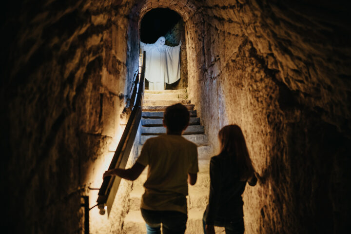 Zwei Kinder gehen in einem schwach beleuchteten Tunnel eine Steintreppe hinauf und stehen oben vor einer Person in einem weißen Laken, die einem Geist ähnelt und die Arme erhoben hat. Die rauen Wände des Tunnels werden von kleinen Lichtern beleuchtet, was eine unheimliche Atmosphäre schafft, die perfekt für ein gruseliges Rahmenprogramm ist.