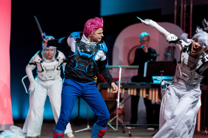 Im Rahmen des Rahmenprogramms 3 treten drei Personen in futuristischen Kostümen auf der Bühne auf. Eine Person mit pinkfarbenen Haaren und einer blauen Hose hält eine Requisitenwaffe in der Hand, während die anderen in weißer Rüstung und Perücken dramatische Gesten machen. Im Hintergrund spielt ein Musiker Percussion-Instrumente und trägt so zur lebhaften Atmosphäre des Rahmenprogramms bei.