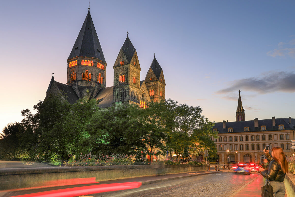 Eine historische Steinkirche mit drei hohen Türmen wird in der Abenddämmerung beleuchtet und bildet den eindrucksvollen Mittelpunkt des abendlichen Rahmenprogramms. Bäume und Autos säumen die Kopfsteinpflasterstraße, und die Lichtspuren vorbeifahrender Fahrzeuge schlängeln sich durch die Szene. Rechts steht eine Person, und im Hintergrund ist ein weiteres Gebäude zu sehen.