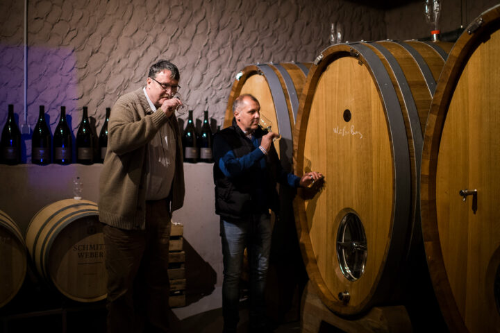 Zwei Männer stehen in einem Keller neben großen Holzfässern und verkosten im Rahmen eines exklusiven Rahmenprogramms Wein aus Gläsern. Im Hintergrund sind Flaschen in Regalen ausgestellt. Die Beleuchtung ist warm und gedämpft.