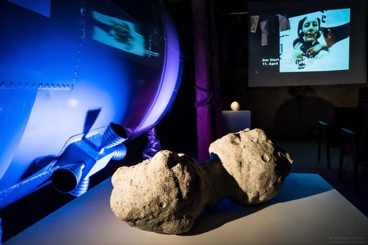 Eine grobe, zweilappige Steinskulptur ruht auf einem weißen Sockel. Links davon befindet sich ein metallisches, blau beleuchtetes Objekt, möglicherweise ein Raumschiff, das an die technologischen Themen anknüpft, die im Rahmenprogramm 3 behandelt werden. Im Hintergrund ist auf einer Projektionsfläche ein Schwarz-Weiß-Bild einer Person mit Text zu sehen.