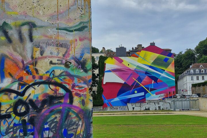 Im Vordergrund ist eine Betonplatte mit bunten Graffiti zu sehen, die den Rahmen für etwas bildet, das Teil eines lebhaften Rahmenprogramms 3 sein könnte. Im Hintergrund ist an einem großen Gebäude ein Wandgemälde mit geometrischen Formen in leuchtenden Farben zu sehen. In der Nähe befinden sich Grasflächen und weitere Gebäude unter einem bewölkten Himmel.